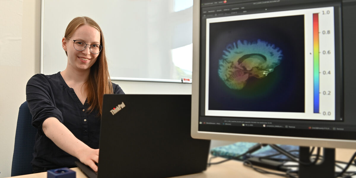 Eine Person sitzt an einem Notebook, auf dem Monitor neben ihr ist ein Gehirnscan zu sehen.