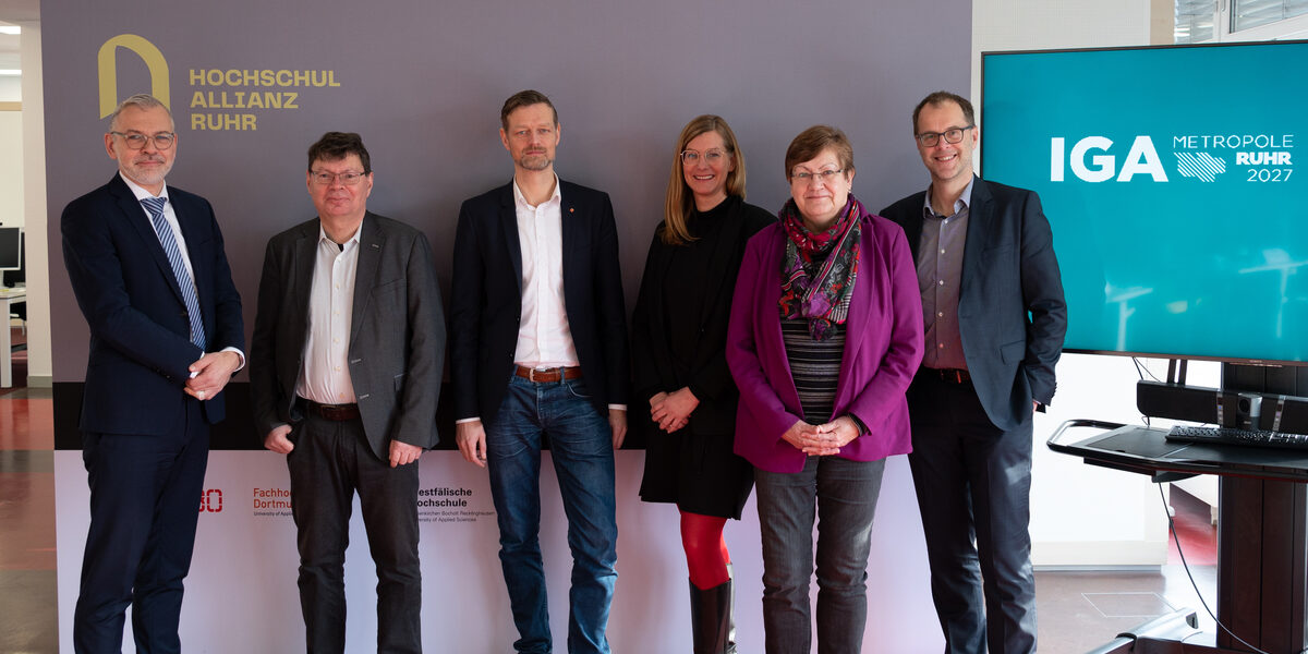 Gruppenfoto mit sechs Personen, die nebeneiner in einer Reihe vor einer Stellwand und einem Display stehen