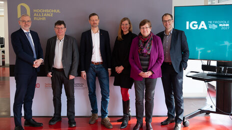 Gruppenfoto mit sechs Personen, die nebeneiner in einer Reihe vor einer Stellwand und einem Display stehen