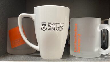 Zwei Tassen mit FH Dortmund in einem Regal mit einer Tasse mit dem Logo der UWA