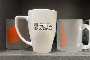 Two mugs with Fachhochschule Dortmund on a shelf with a mug bearing the UWA logo