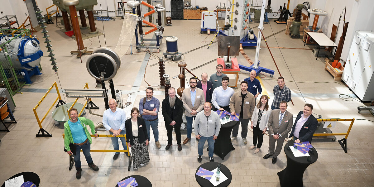 16 people, photographed from an elevated position, stand in a high hall surrounded by technical equipment and a few black-clad bar tables with brochures on them.