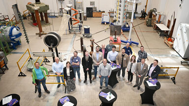 16 people, photographed from an elevated position, stand in a high hall surrounded by technical equipment and a few black-clad bar tables with brochures on them.