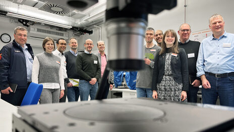 Elf Personen stehen in einem Raum mit dem Gesicht zur Kamera. In der Mitte im Vordergrund ragt von oben, ganz nah und unscharf, ein silberner Zylinder ins Bild. Im unteren Vordergrund, ebenso nah, ist eine dunkle Platte zu sehen.