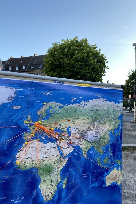 Deutschland ist auf einer Weltkarte durch eine Stecknadel markiert. Orangefarbene Fäden führen von hier aus zu anderen Ländern.