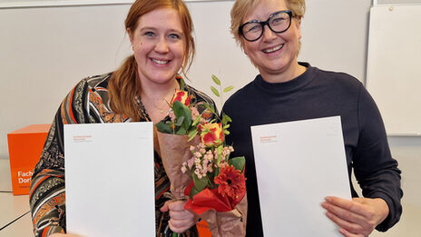 Two people are standing next to each other, each holding a bouquet of flowers and a white folder in front of them.