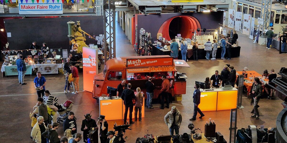 In einer großen Halle mit vielen Menschen steht in der Mitte ein orangefarbener Oldtimer mit Messeaufbauten.