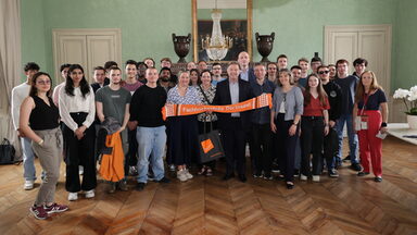A group of people are standing in a hall, the people in the middle are holding an orange scarf with the words Fachhochschule Dortmund written on it.