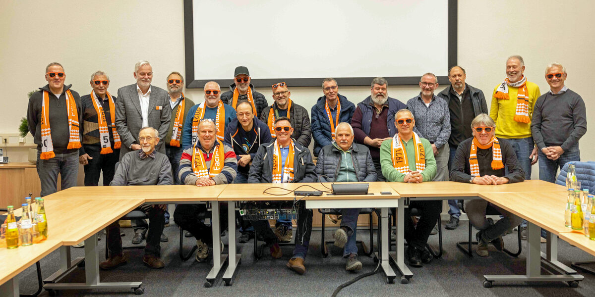Eine große Personengruppe in einem Seminarraum. Viele von ihnen tragen orangefarbene Sonnenbrillen der FH Dortmund.