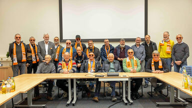 Eine große Personengruppe in einem Seminarraum. Viele von ihnen tragen orangefarbene Sonnenbrillen der FH Dortmund.