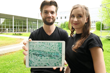 Zwei Personen sitzen auf dem Campus der FH Dortmund. Sie halten ein Tablet in die Kamera. Darauf ist ein architektonischer Entwurf zu sehen.