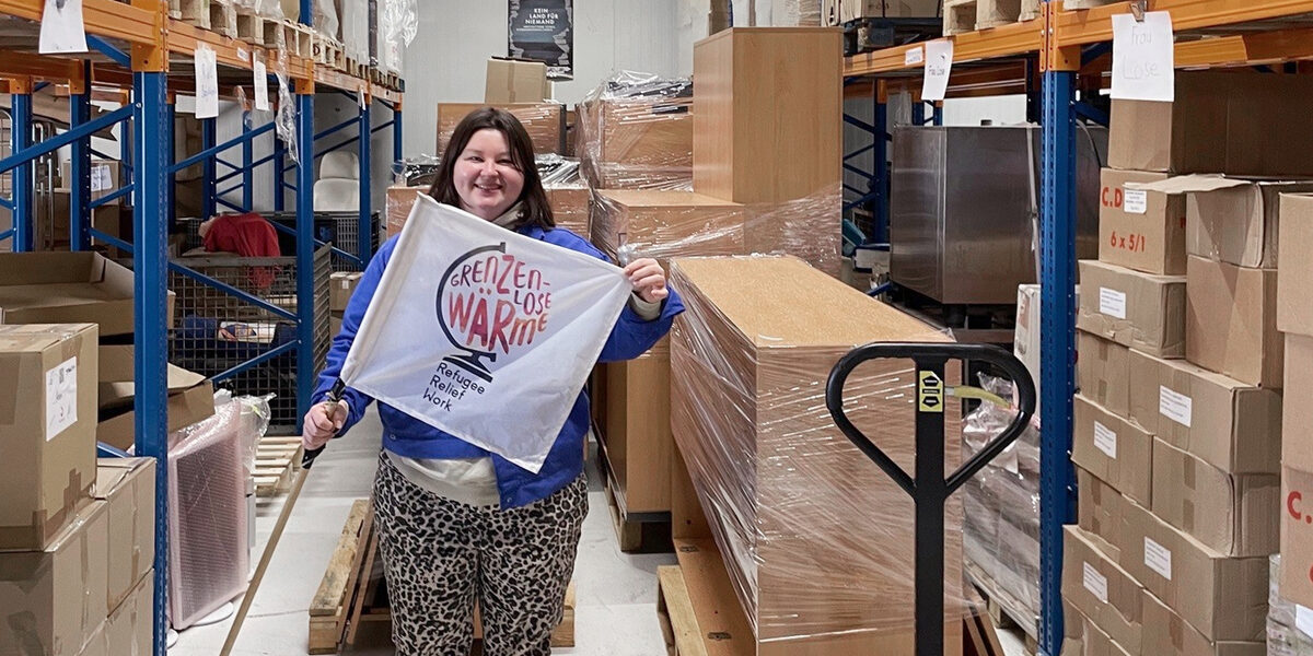 Eine Person hält eine Fahne mit dem Schriftzug "Grenzenlose Wärme" in den Händen. Die Person steht in einem Lager zwischen hohen Regalen, neben ihr ein Hubwagen mit in Folie gewickelten Möbeln.
