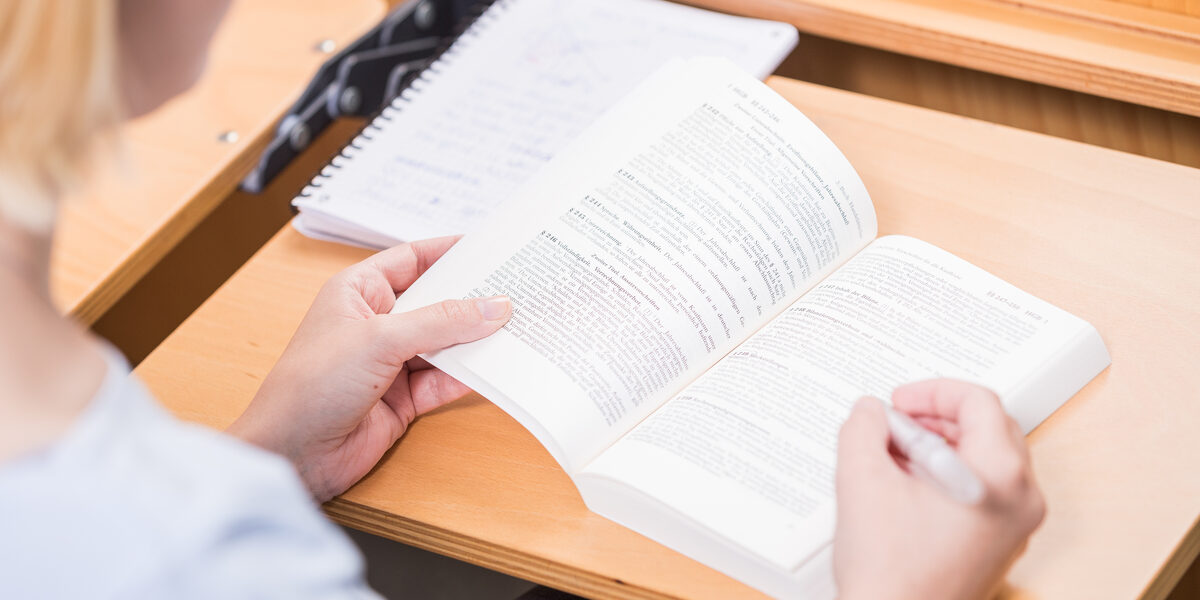 Foto über die Schulter einer Studentin an einem Tisch im Hörsaal. Sie schlägt das Handelsgesetzbuch auf.