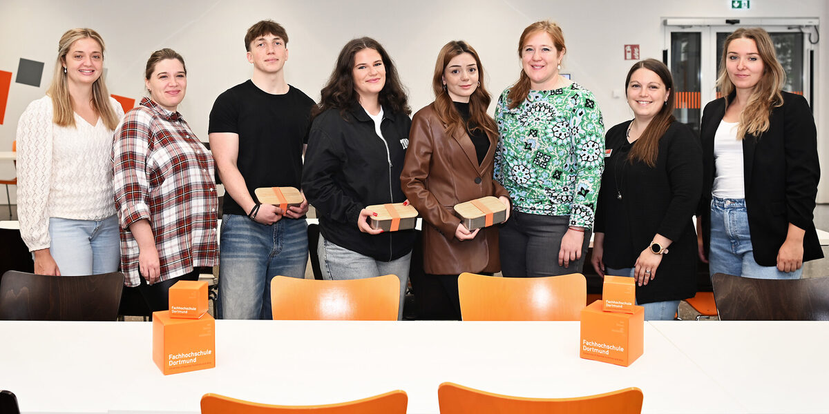 Acht Personen stehen in einem hellen, großen Raum hinter einem weißen Tisch und schauen freundlich in die Kamera.