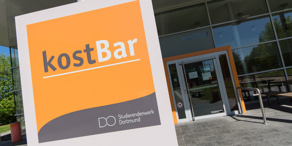 Photo of a sign with the logo of the kostBar canteen, in the background the entrance to the kostBar.