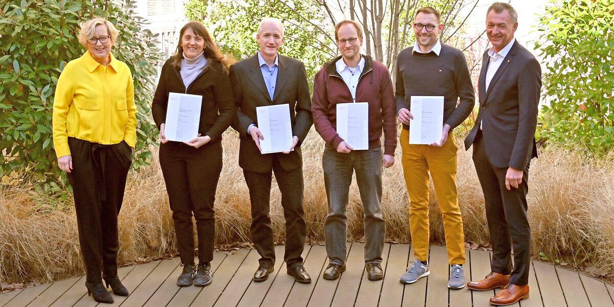 Six people stand next to each other in front of a planted house facade, the four people in the middle hold a document in front of them.