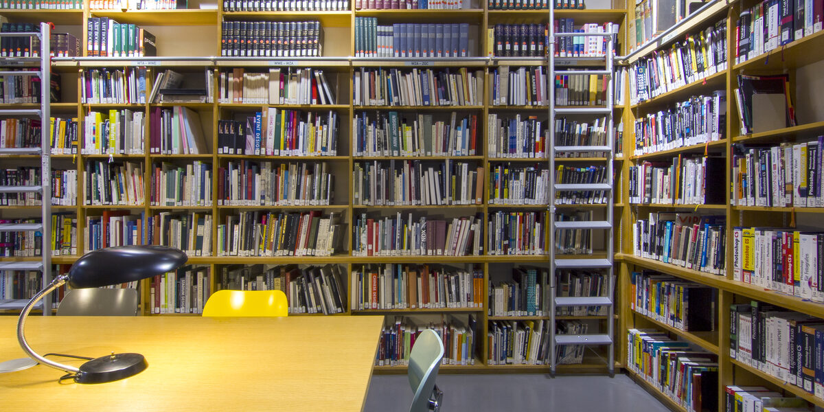 Foto des Lesesaals in der Bibliothek mit Bücherregalen, Tischen, Leselampen und Stühlen__Photo of the library reading room with book shelves, desks, reading lamps and chairs