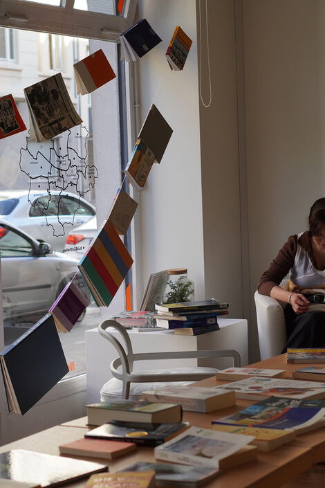 Books are hanging on a string, on the right a person is sitting and reading a book.