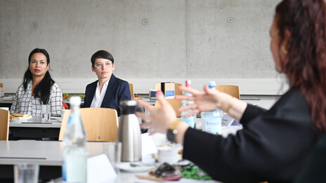 Rechts im Vordergrund sitzt eine Person, zu sehen von schräg hinten, und gestikuliert in Richtung zweier Personen, die, von vorn gesehen, im Mittelgrund in der linken Bildhälfte sitzen. Sie alle sitzen an weißen Tischen, hinten schließt eine graue Betonwand den Raum ab.