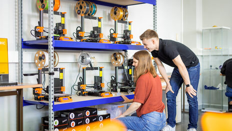Foto von zwei Studierenden, die am 3D-Drucker-Regal stehen/hocken.