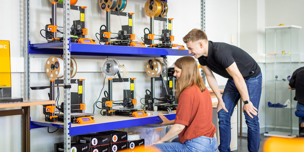 Foto von zwei Studierenden, die am 3D-Drucker-Regal stehen/hocken.