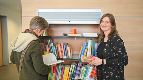 Zwei Frauen stehen vor einem Bücherregal. Die Frau auf der linken Seite steht mit dem Rücken zur Kamera und liest in einem Buch. Die Frau auf der rechten Seite hält einen Stapel Bücher in den Händen und lächelt in die Kamera.__ Two women are standing in front of a book shelf. The woman on the right has her back turned to the camera and is reading a book. The woman on the right is holding a pile of books and smiling at the camera.