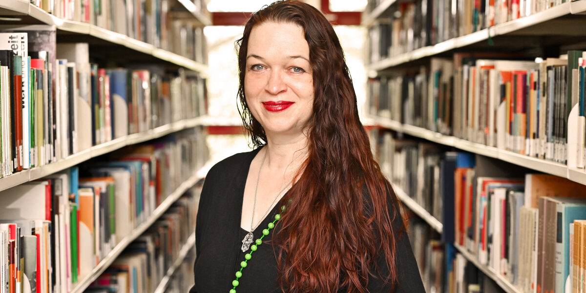 A person in a black top and with long dark red hair stands between bookshelves and looks into the camera.