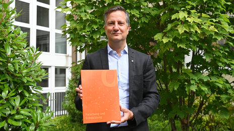 Ein Mann hält mit beiden Händen ein orangefarbenes Magazin vor sich. Im Hintergrund ist ein begrünter Innenhof zu sehen.
