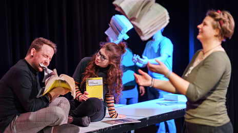 Foto von Schauspieler:innen, die eine Szene im Theaterlabor performen und dabei mit Büchern jonglieren.
