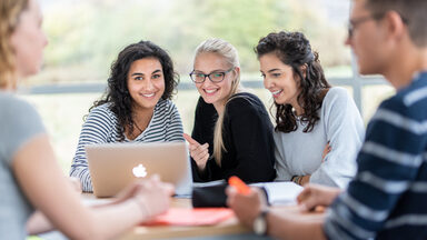 Photo of five students at a group table. The focus is on three female students sitting next to each other, with a laptop on the table in front of them.