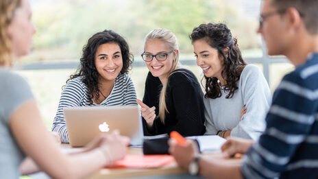 Photo of five students at a group table. The focus is on three female students sitting next to each other, with a laptop on the table in front of them.