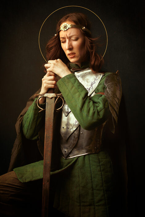 A person with long red hair against a black background in a knight's armor is leaning on a sword, keeping his eyes closed and his forehead wrinkled.