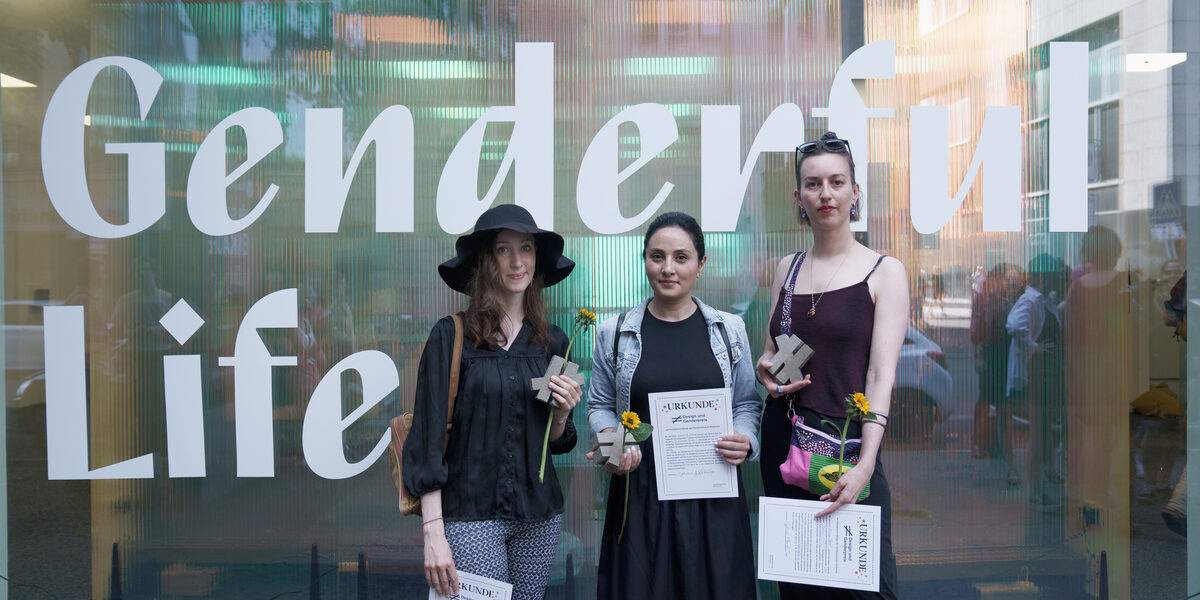 Three people stand in front of a semi-transparent shop window with the words "Genderful Life". They are each holding a certificate, a sunflower and a gray inequality sign.