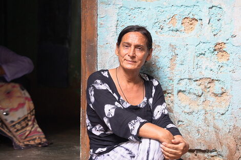 A person in light, summery clothing sits in front of a house entrance and smiles gently into the camera.