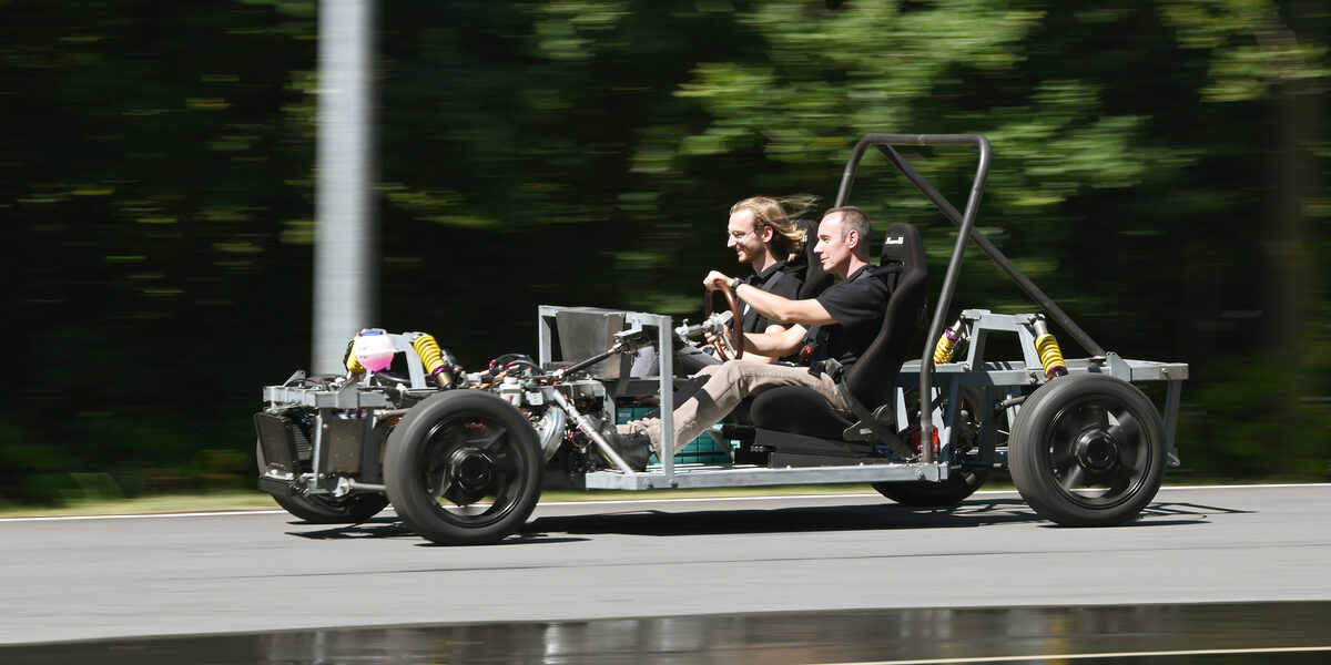 Zwei Männer fahren in einem offenen, unverkleideten Fahrzeug über eine Straße.