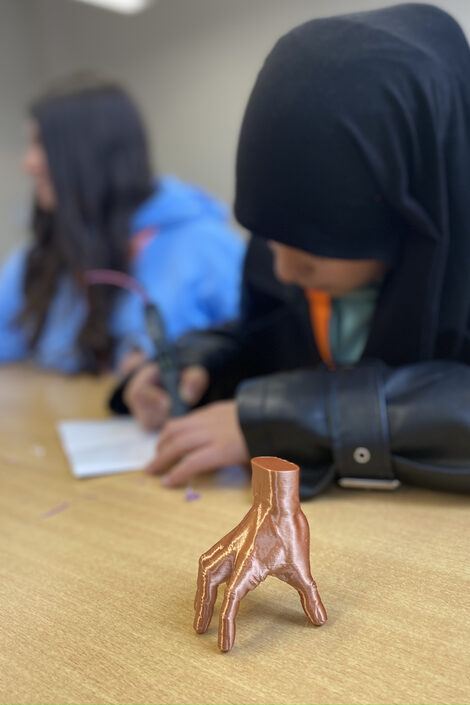 A model of a hand stands on a table, with two people sitting behind it, one of whom is holding a pen over a sheet of paper.