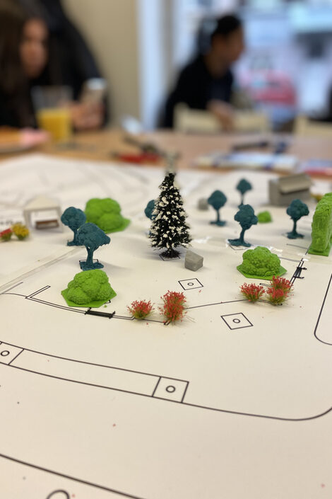 Im Vordergrund ist ein einfacher Lageplan zu sehen mit Modellbäumen, die darauf stehen. Der unscharfe Hintergrund zeigt Personen, die am Tisch sitzen.