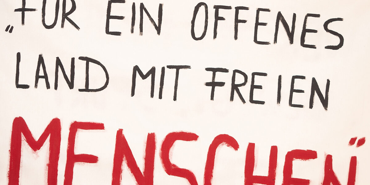 Plakat mit Aufschrift: Für ein offenes Land mit freien Menschen. Im Hintergrund andere im Bildanschnitt abgeschnittene Plakate mit Auschrift.