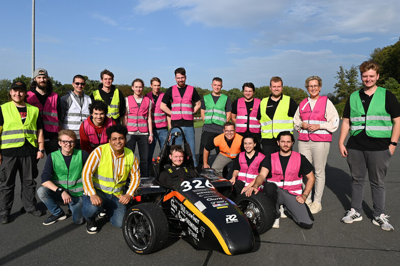 A large group of people stand around the Fachhochschule Dortmund racing car.