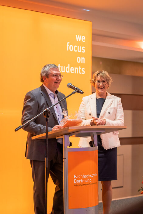 Two people are standing on a stage at a lectern.