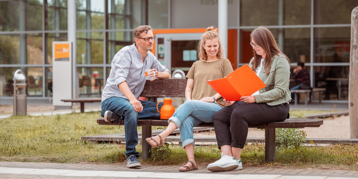 Photo of 3 people sitting on a wooden bench outside the kostBar and talking.