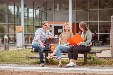 Photo of 3 people sitting on a wooden bench outside the kostBar and talking.