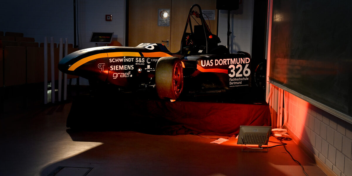 A racing car is dramatically illuminated on a pedestal in a lecture hall.