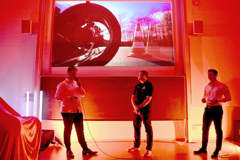 Three people on a red-lit stage under a video beamed onto the wall.