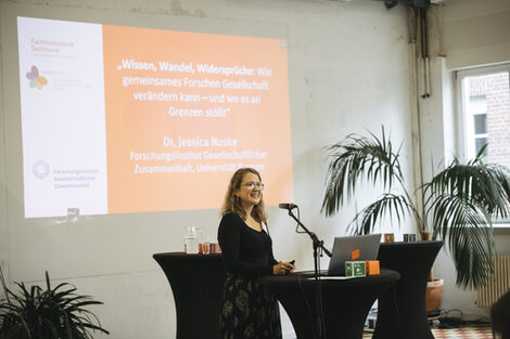 Eine Person steht an einem Stehtisch und spricht in ein Mikro. An der Wand hinter ihr eine Präsentation