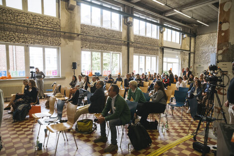 Blick durch eine Halle auf ein auf Stühlen sitzendes Publikum.
