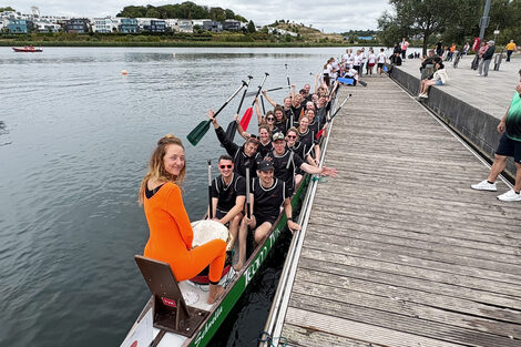 View from the jetty into a narrow boat with 18 people in black jerseys and paddles in their hands. At the very front sits a 19th person in orange.
