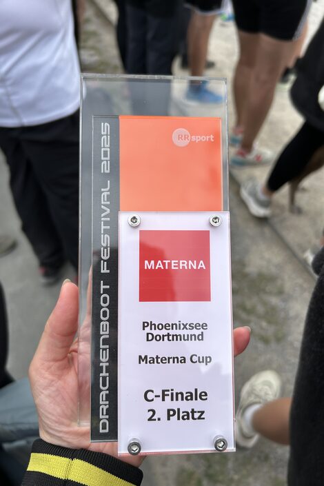 One hand holds a trophy made of Plexiglas and plastic with the inscription "Materna. Phoenixsee Dortmund. Materna Cup. C-final, 2nd place". The legs of several people can be seen around it.