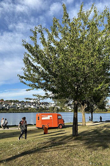 An orange vintage car is parked on the shore of a lake under a blue sky. In the foreground, a tree rises into the sky, some people walk along the shore.