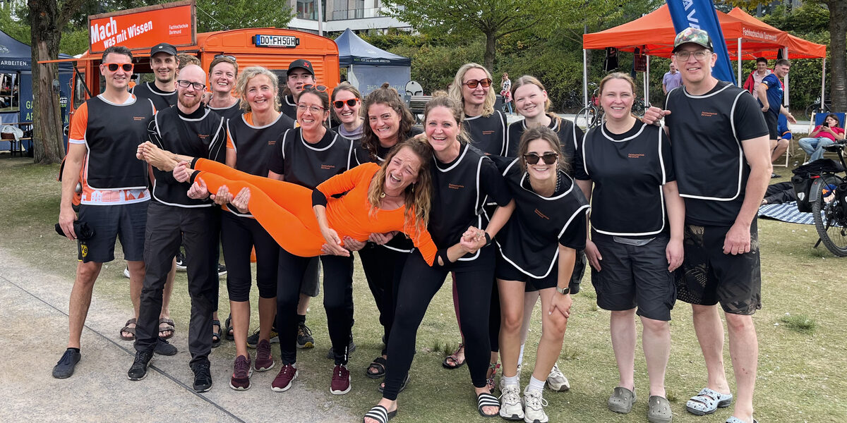 15 people in black jerseys carry another person in orange on their hands, stand next to each other and look into the camera. In the background, a car and two tents, both orange, stand on a meadow.